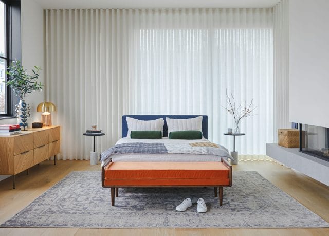 A bedroom is shown with a bright orange bench, neutral rug, and blue bedding.