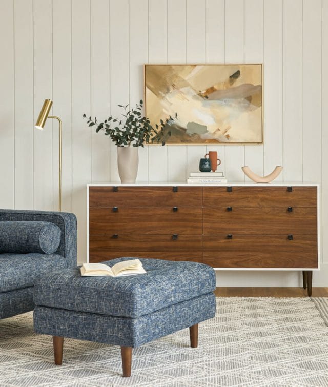 A white and brown wood 6-drawer dresser in a modern living room next to a blue chair and ottoman. 