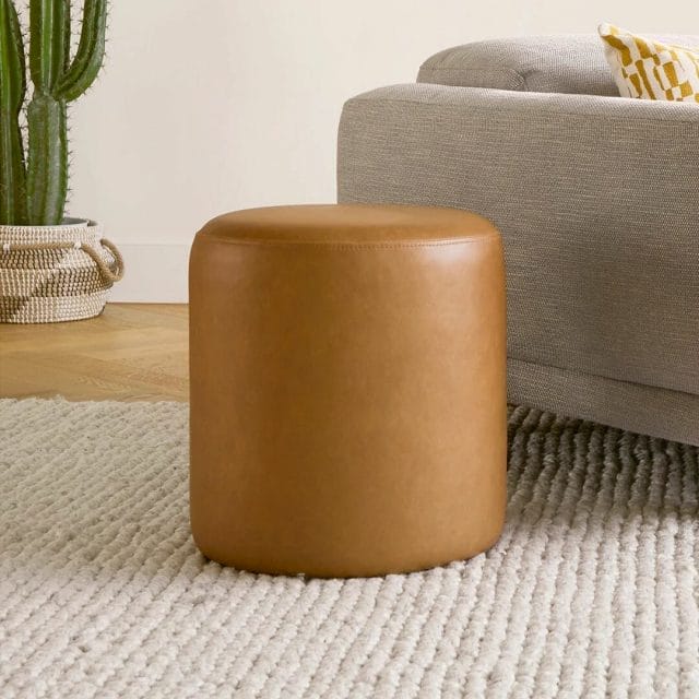 A brown leather ottoman next to a gray sofa on a white rug. 
